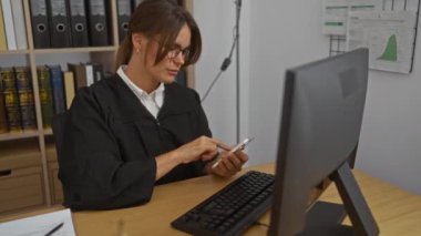 Mahkeme salonunda çalışan bir kadın akıllı telefon ve bilgisayar kullanırken aynı zamanda yargıç cübbesi ve gözlüğü takıyor ve rafta yasal kitaplar var.