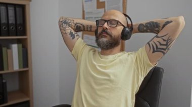 Sakallı, dövmeli, gözlüklü ve kulaklıklı İspanyol bir adam ofiste dinleniyor.