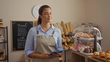 Kapalı bir pastanede elinde tabletle ayakta duran genç bir kadın, dükkânın etrafında çeşitli hamur işleri ve ekmek somunları sergileniyor.