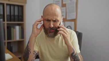 Ofiste sakallı İspanyol bir adam telefonda konuşuyor. Stresli ve kafası karışık görünüyor. Üzerinde sarı bir gömlek, görünür dövmeler var.