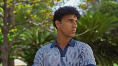 Arka planda ağaçlarla dolu şehir parkında genç bir adam, kollarını kavuşturmuş, günlük gömlekler giyiyor.