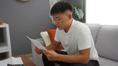 Oturma odasında oturan genç adam, düşünceli bir ifadeyle evrak okuyor, beyaz gömlekli bir kanepede oturuyor, rahat bir evin içi ile çevrili.