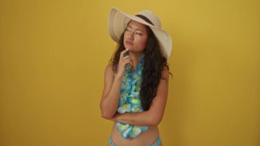 Yaz şapkalı genç Çinli kadın, Hawaii lei 'li düşünceli, düşünceli ve ciddi görünümlü, el ele tutuşmuş, sarı arka plandaki bir fikri ya da soruyu düşünüyor.