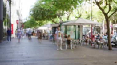Güneşli bir sokakta yürüyen insanlar kafeleri, ağaçları ve şehir merkezinde bulanık bir geçmişi olan insanlar.