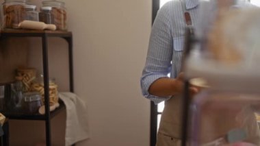 Taze ekmek somunları ve raflardaki kurabiye kavanozlarıyla çevrili, rahat bir fırın ortamında renkli bardaklarla tepsiyi tutan bir kadın.