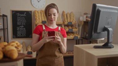 Kırmızı gömlekli ve kahverengi önlüklü bir fırıncı, arka planda ekmek ve hamur işleri sergilenen bir fırının arkasında dururken akıllı telefon kullanıyor..