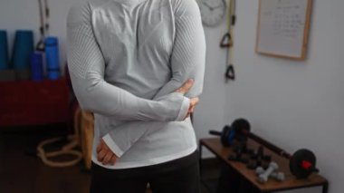 Spor salonundaki adam dirseğini tutuyor. Arka planda spor aletleri ve spor salonu seti var.