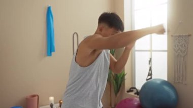 Boks hareketleri yapan genç bir adam, arka planda spor topları ve kapalı bitkilerle dolu iyi aydınlatılmış bir spa odasında sağlık ve sağlığa odaklandığını gösteriyor.