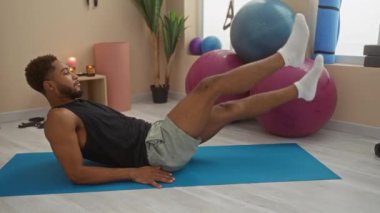 Spor salonundaki yoga minderinde egzersiz yapan genç bir adam. Arka planda spor aletleri var.