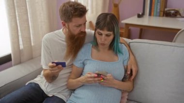 Akıllı telefon ve kredi kartı kullanan hamile bir çift rahat bir oturma odasında oturuyor.