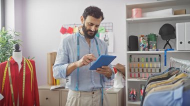 Bir atölyede sakallı bir terzi mankenler, kumaşlar ve renkli iplikler arasında tablet kullanıyor..