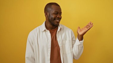 Young man extends hand in welcoming gesture against isolated yellow background, showcasing casual attire and friendly demeanor. clipart