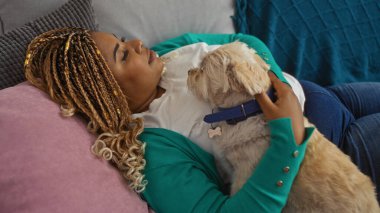 Genç Afro-Amerikalı kadın, örgülü köpeğiyle oturma odasında kanepede dinleniyor, ev ortamında rahat bir ortam sergiliyor..