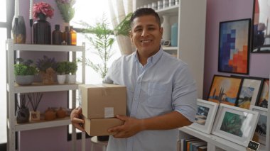 Çiçekler ve sanat eserleriyle süslenmiş bir fırın odasında paketleri tutan genç adam, dostça bir gülümseme sergiliyor.