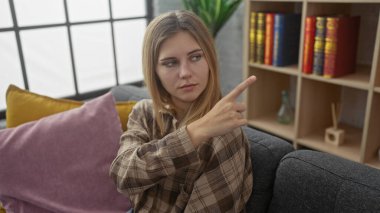 Sarışın kadın, rahat, modern bir oturma odasından kitap, yastık ve kanepeyi işaret ediyor..