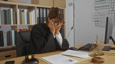 Kadın yargıç, mahkemeyle ilgili materyallerle çevriliyken hukuk kitapları ve belgelerle dolu bir ofiste masasının başında stresli bir şekilde duruyor..