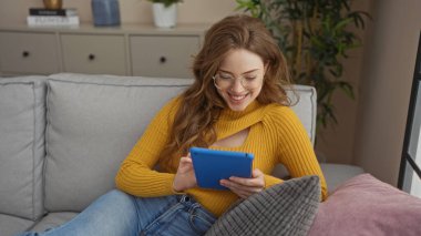 Güzel, genç, sarışın, gözlüklü ve sarı kazaklı bir kadın kanepede oturuyor, gülümsüyor ve rahat bir oturma odasında mavi bir tablet kullanıyor..