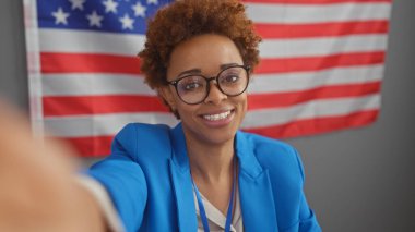 Gözlüklü ve mavi ceketli gülümseyen Afrikalı Amerikalı bir kadın arka planda Amerikan bayrağıyla selfie çekiyor..