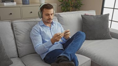 Oturma odasındaki koltukta oturan sakallı, kulaklık takan ve sıradan bir şekilde telefonuna bakan genç İspanyol bir adam..