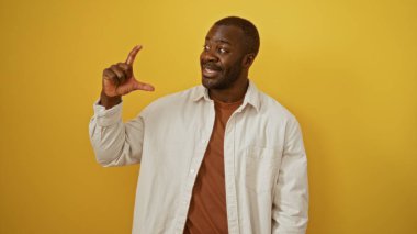 İzole edilmiş sarı arka planda büyük bir jest yapan genç adam, parmaklarıyla ve neşeli bir ifadeyle bir konsepti ifade ediyor, gündelik tarzını canlı renklerle harmanlıyor..