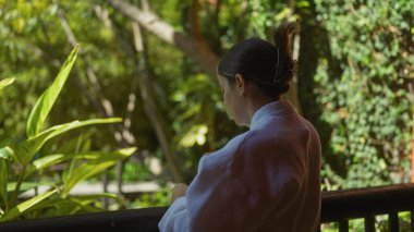 Young woman relaxing on a shaded terrace surrounded by tropical plants in bali, indonesia clipart