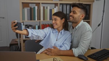 Kadın ve erkek bir ofiste selfie çekiyorlar. Birbirlerine gülümsüyorlar ve yan yana oturuyorlar. İş birliğini ve iş ortaklığını profesyonel bir iç mekan ortamında somutlaştırıyorlar.