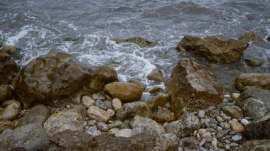 Sahilde kayalara vuran dalgalı kıyı şeridi, ıslak taşlar ve çalkantılı suyla doğal kıyı manzarasını gözler önüne seren elementler engebeli güzelliği ve doğal dokuları vurguluyor..