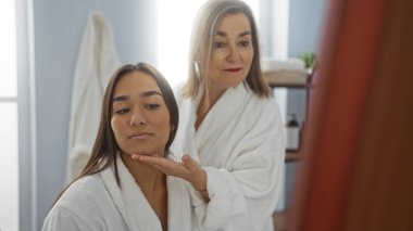 Beyaz bornoz giyen anne ve kız spa ortamında birlikte iyi vakit geçirmenin keyfini çıkarıyorlar. Aynaya yansıyor ve iç mekanda huzur veriyor..