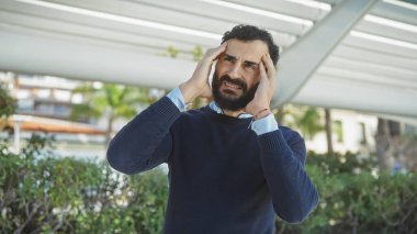 Sakallı, sıkıntılı orta yaşlı bir adam parkta dikiliyor, hayal kırıklığı ya da baş ağrısı içinde şakaklarına dokunuyor..