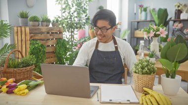 Çiçekçide dizüstü bilgisayar kullanan önlüklü gülümseyen Asyalı adam. Etrafı çiçek ve bitkilerle çevrili..
