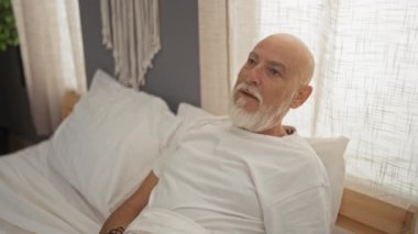 Kel ve sakallı bir adam rahat bir yatak odasında dinleniyor. Pencereden süzülen güneş ışığının tadını çıkarırken bir yatakta oturuyor..