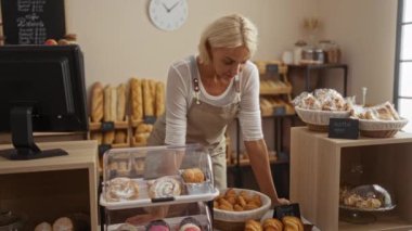 Bir pastanedeki sarışın kadın tezgahın arkasında duruyor. Etrafı taze hamur işleri ve fırınlarla çevrili. Bu da hoş ve hoş bir iş ortamı olduğunu gösteriyor.