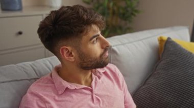 Pembe polo tişörtlü, sakallı genç adam, rahat bir oturma odasında oturur, kameraya bakar hafif bir gülümsemeyle, rahat ve rahat bir atmosfer sergiler.
