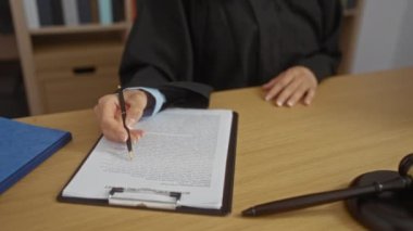 Kadın yargıç, masa başında tokmak ve arka planda kitap raflarıyla ofiste yasal belgeleri imzalıyor.