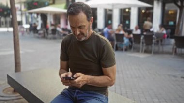 Latin kökenli orta yaşlı bir adam arka planda insanların ve restoranların olduğu bir kentteki bir bankta akıllı telefon kullanırken kapalı yumruklarla kutlama yapıyor..