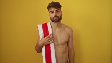 Plaj havlusunu tutan İspanyol asıllı genç adam el hareketini durduruyor. Ciddi bir ifadeyle, izole edilmiş sarı arka planda savunmaya güveniyor.