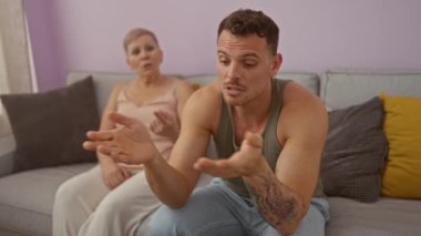 Dövmeli bir adam ve oturma odasındaki koltukta oturan yaşlı bir kadın gri yastıklı ve mor duvarlı ciddi bir tartışma içerisindeler.