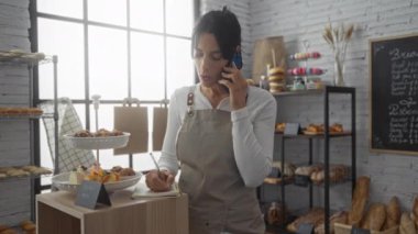 Bir pastanede arka planda hamur işleri ve ekmeklerle defterde yazan bir kadın telefonda konuşuyor.