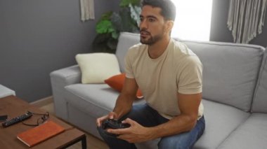 Modern bir oturma odasında video oyunu oynayan sakallı genç adam, ekranda odak ve konsantrasyon gösteriyor, rahat yastıklar ve ev bitkileriyle çevrili..