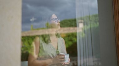 Camdan dışarı bakan bir kadın, elinde bir bardak tutarken yansımaları olan sessiz bir kafe.