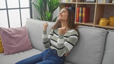 Neşeli, genç İspanyol bir kadın, rahat bir oturma odasında gri bir kanepede oturmuş zaferini kutluyor..