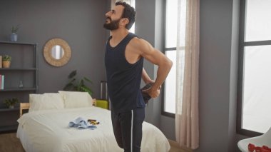 Sakallı bir adam modern bir yatak odasında gün ışığı ve günlük kıyafetlerle esniyor..