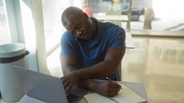 Ofiste çalışan bir adam iş yerinde bilgisayarını kullanarak telefonla konuşurken notlar yazıyor.