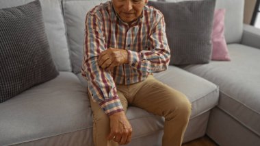 Orta yaşlı bir adam, kanepede rahat bir oturma odasında, ekose gömlek ve bej pantolon giymiş bir şekilde otururken kolunu kaşıyordu..