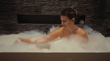 Huzurlu genç bir kadın, lüks bir banyonun zarif taş dekoruyla çevrili köpük banyosunda dinleniyor..