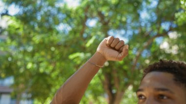 Genç adam güneşli bir günde şehir parkında ağaçların altında yumruk kaldırıyor.