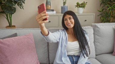 Uzun esmer saçlı, genç, çekici bir İspanyol kadın şık dekore edilmiş bir oturma odasında rahat bir koltukta otururken selfie çekiyor..