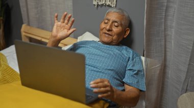 Gri saçlı yaşlı İspanyol bir adam, yatak odasından bir video çağrısı alıyor, bir dizüstü bilgisayar kullanıyor ve neşeli bir ifadeyle el sallıyor..