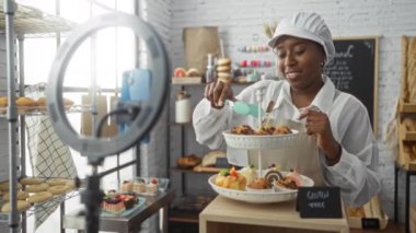 Kadın pastanede, arka planda bir halka ışığı ve fırınlanmış malların rafları olan katmanlı bir tezgahta glütensiz hamur işleri düzenleyen bir video kaydediyor.