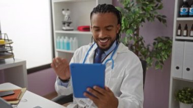 Örgü ve steteskop takan genç adam tabletle yapılan bir video görüşmesi sırasında gülümsüyor ve modern bir klinikte bitki ve tıbbi malzemelerle birlikte oturuyor..